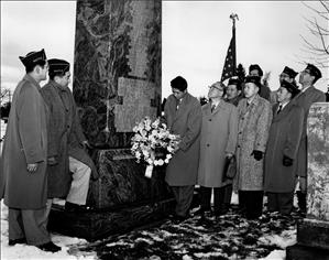 A group of Asian men in long winter coats stand at the base of a marble monument. One of them places a wreath of flowers on the monument. There is snow on the ground. 